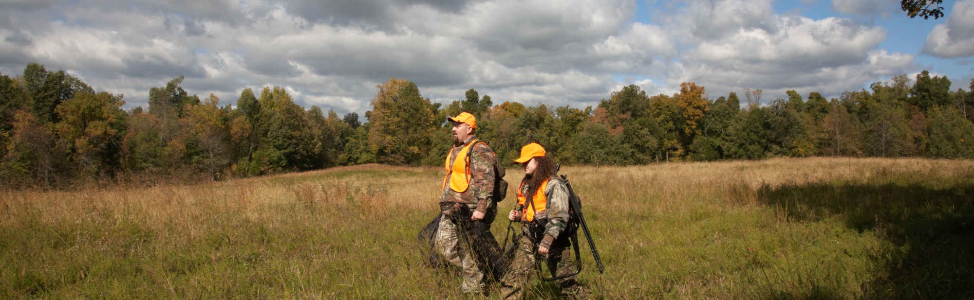 Two hunters walking in a grassy field surrounded by trees.